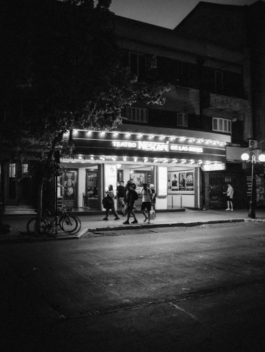 A black and white street scene with people outside a lit storefront at night.